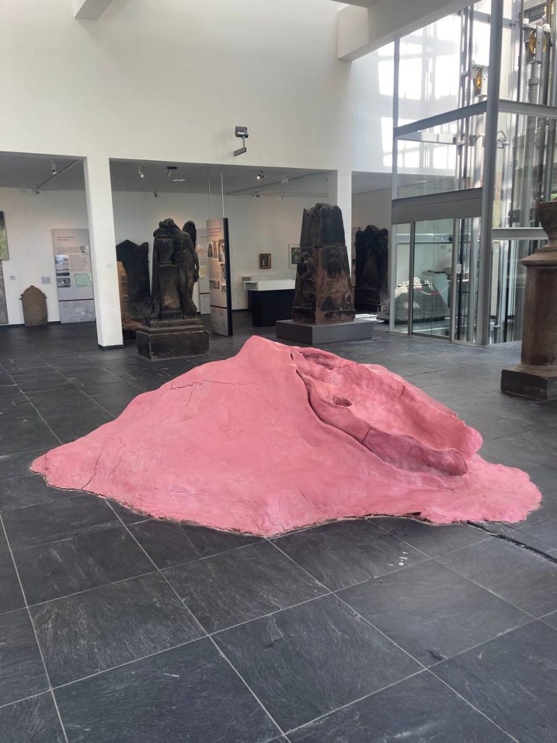 Arbeit Spewing Humus im Sepulkralmuseum Kassel. Gesamtansicht. Pinker Ton in dem eine Vagina eingearbeitet ist. Aus ihr spritzt Wasser.