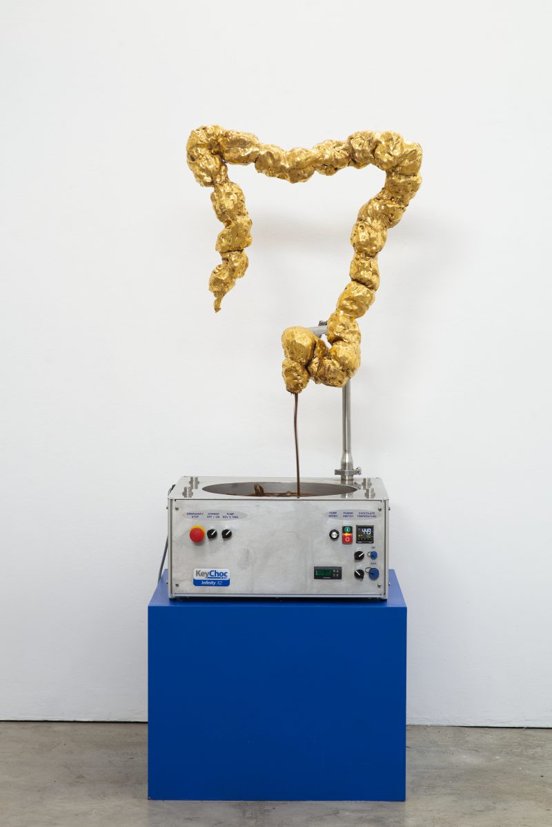 Goldener Dickdarm aus Keramik. Handgeformte Skulptur von der Künstlerin Laura Pientka. Der Dickdarm pumpt warme Schokolade aus seinem Ende. Dies lässt an Exkremente erinnern